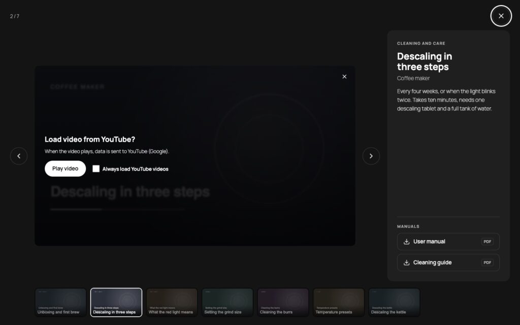 Full-screen view on a dark stage with the consent panel Load video from YouTube over the preview image, previous and next arrows, a counter reading 2 of 7, the title Descaling in three steps with its description and product, two manual download buttons and a strip of all seven videos below.